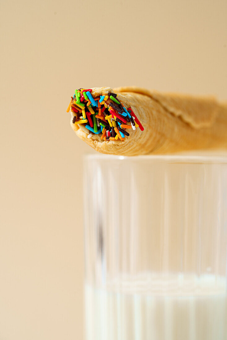 Close-up image of a waffle roll topped with colorful sprinkles resting on the rim of a glass of milk against a beige background.