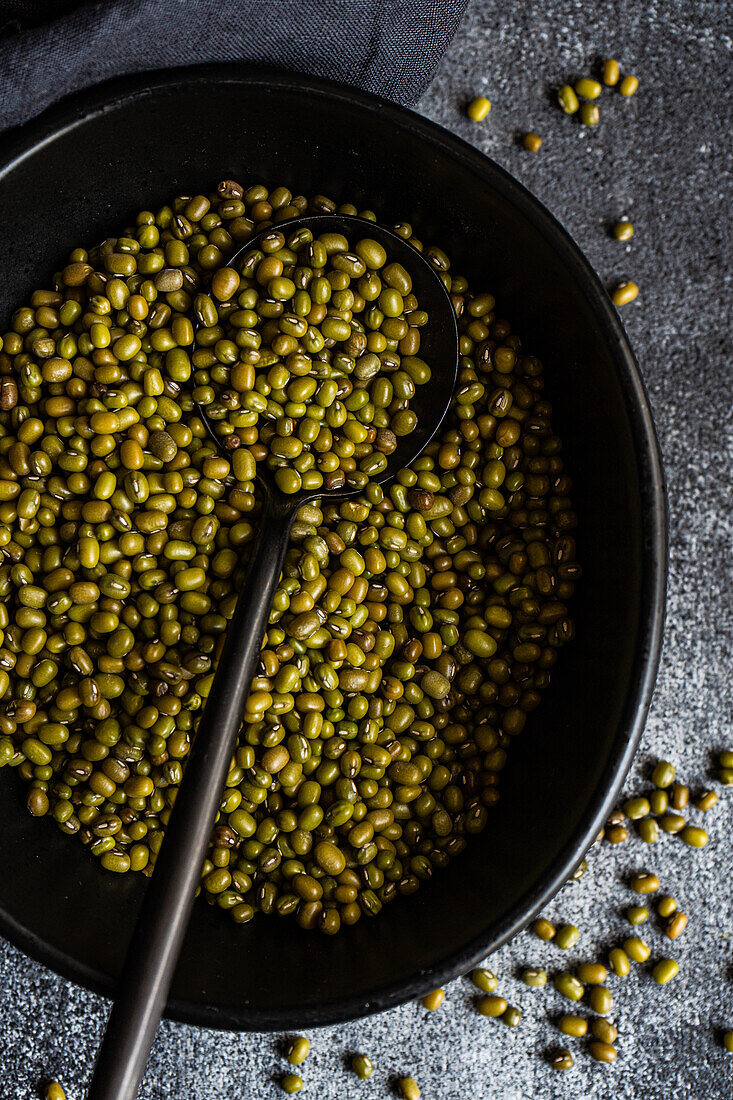 A rustic display of raw organic mung beans heaped in a dark skillet, with a black spoon partially filled