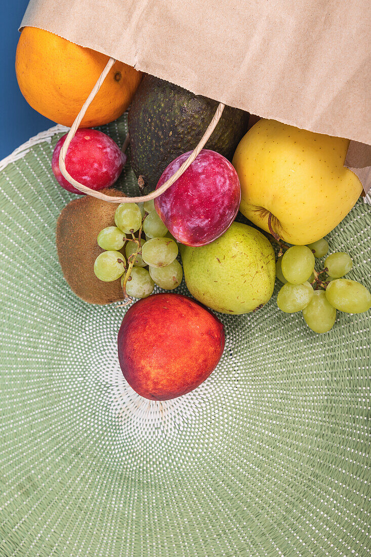 Top view of a vibrant selection of fresh fruits spilling from a paper bag onto a textured green mat. Includes oranges, avocados, apples, kiwis, and grapes.