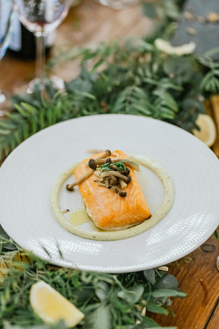 Perfekt gegartes Lachsfilet mit zarten Pilzen, serviert auf einem weißen Teller mit Kräuterpüree, inmitten einer festlich gedeckten Tafel