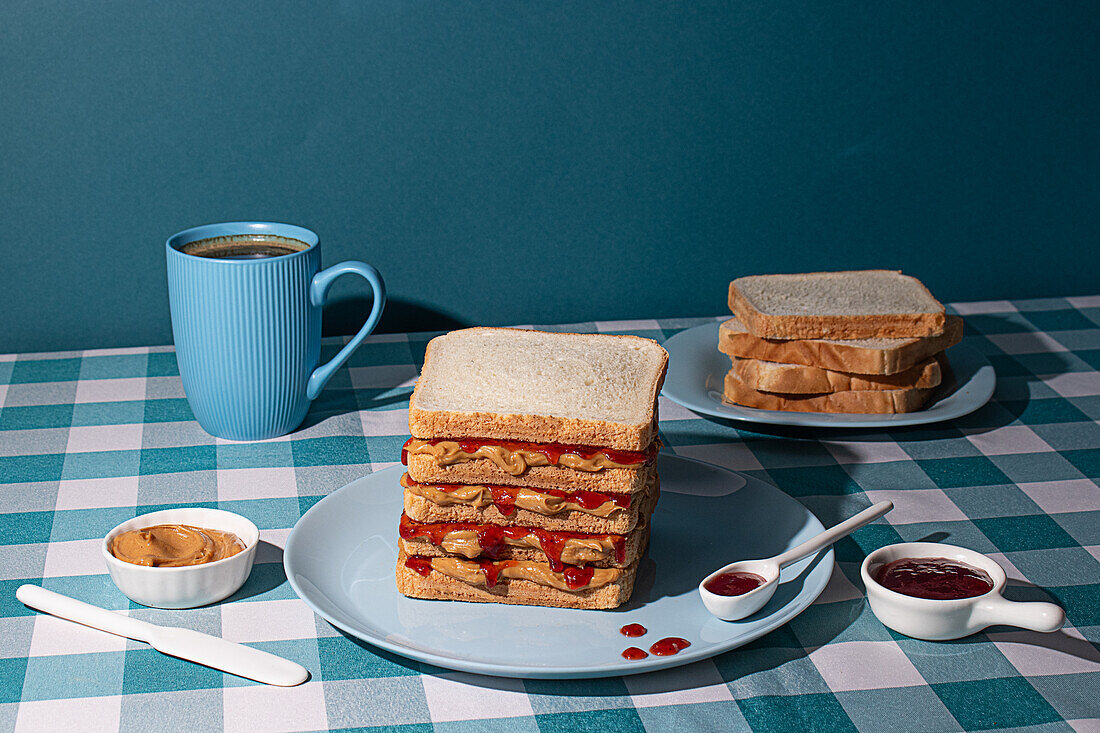 Stapel Erdnussbutter-Gelee-Sandwiches auf einem blauen Teller, mit einer Tasse amerikanischem Kaffee und einigen Brotscheiben auf blauem Hintergrund