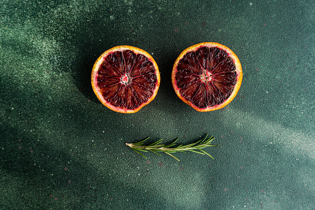 Two halves of juicy blood oranges with a sprig of rosemary on a textured green background