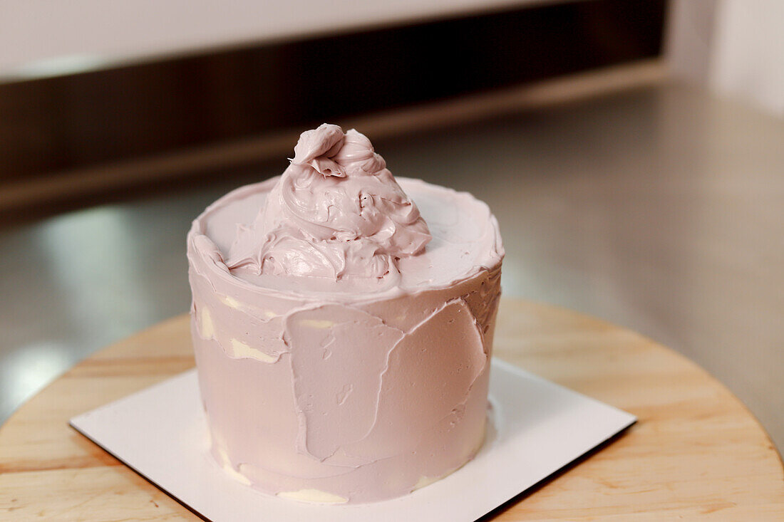 A single-layer cake with pink frosting being applied on a wooden surface, indicative of a baking process in a home kitchen