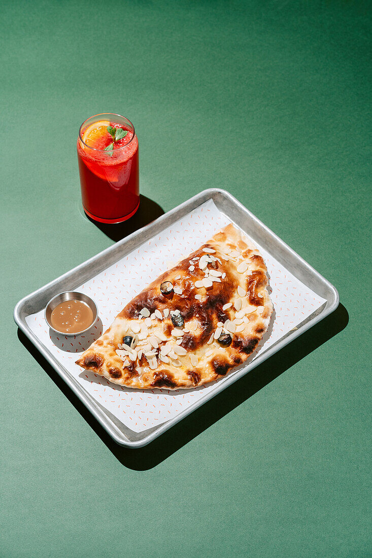 Goldener gebackener Cheburek neben einem süßen gebackenen Cheburek mit Mandeln, begleitet von einer erfrischenden Beeren-Orangen-Limonade vor einem grünen Hintergrund.