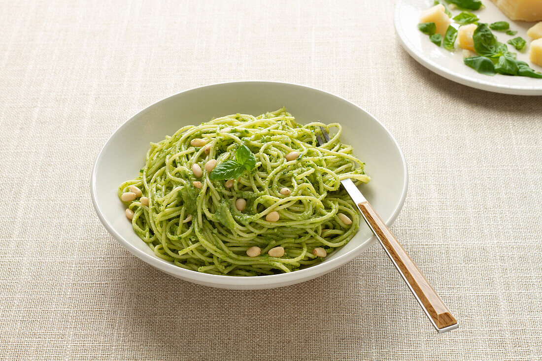 A white bowl filled with spaghetti pesto topped with pine nuts and a basil leaf, accompanied by a fork on a beige background.