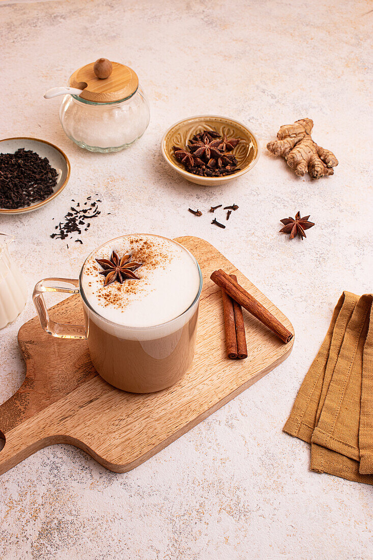 From above, in rustic style a frothy chai latte with star anise on a wooden board surrounded by cinnamon sticks, fresh ginger, tea leaves, a milk jar and a brown napkin