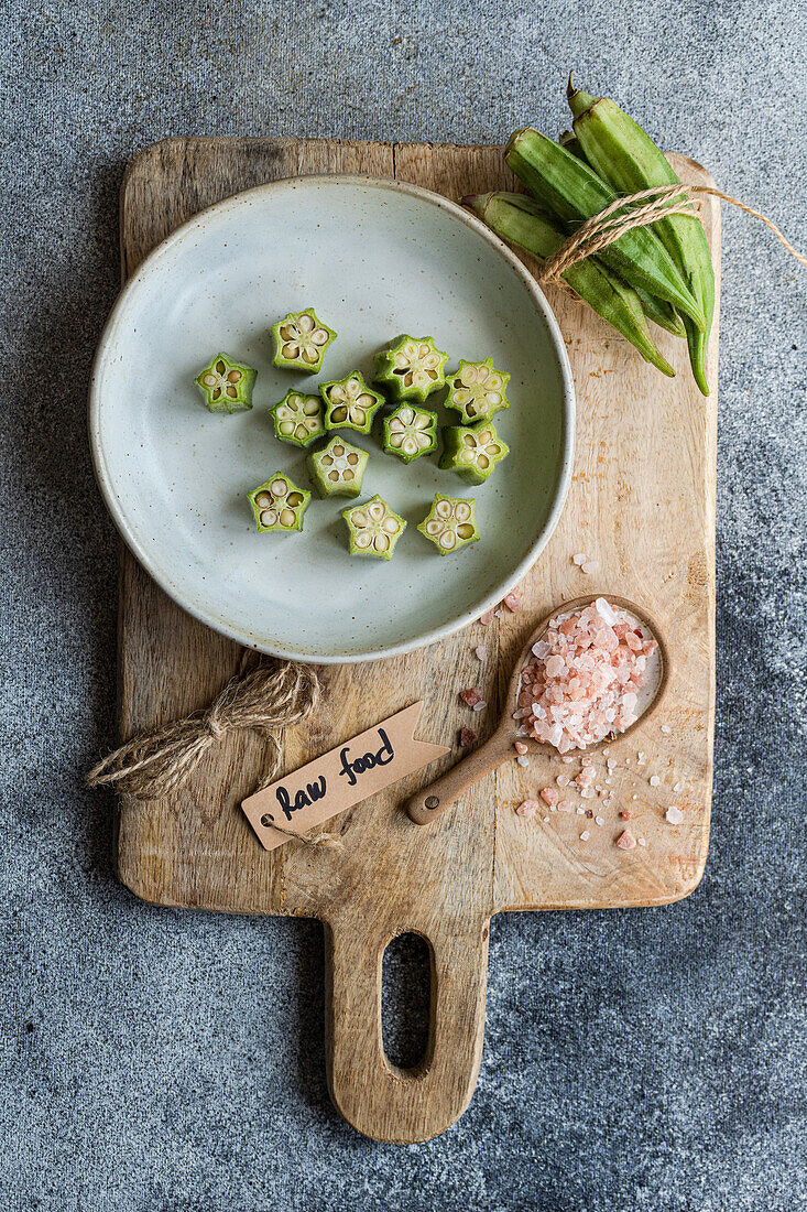 Raw, sliced bamia displayed on a rustic wooden board, accompanied by a raw food label, himalayan pink salt, and tied okra, ideal for a Mediterranean cuisine theme