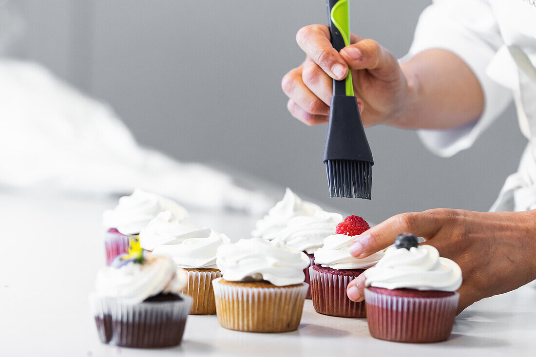 Unbekannte Bäckerin mit Silikonpinsel, die süßen Sirup auf Beeren verteilt, während sie vegane Cupcakes in der Küche zubereitet