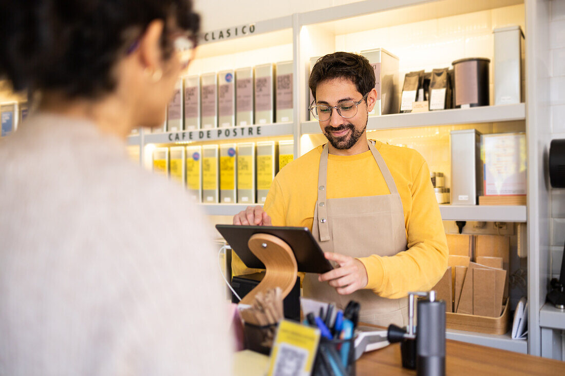 A cheerful barista in a yellow sweater accepts a mobile contactless payment from a customer at a cozy café counter.