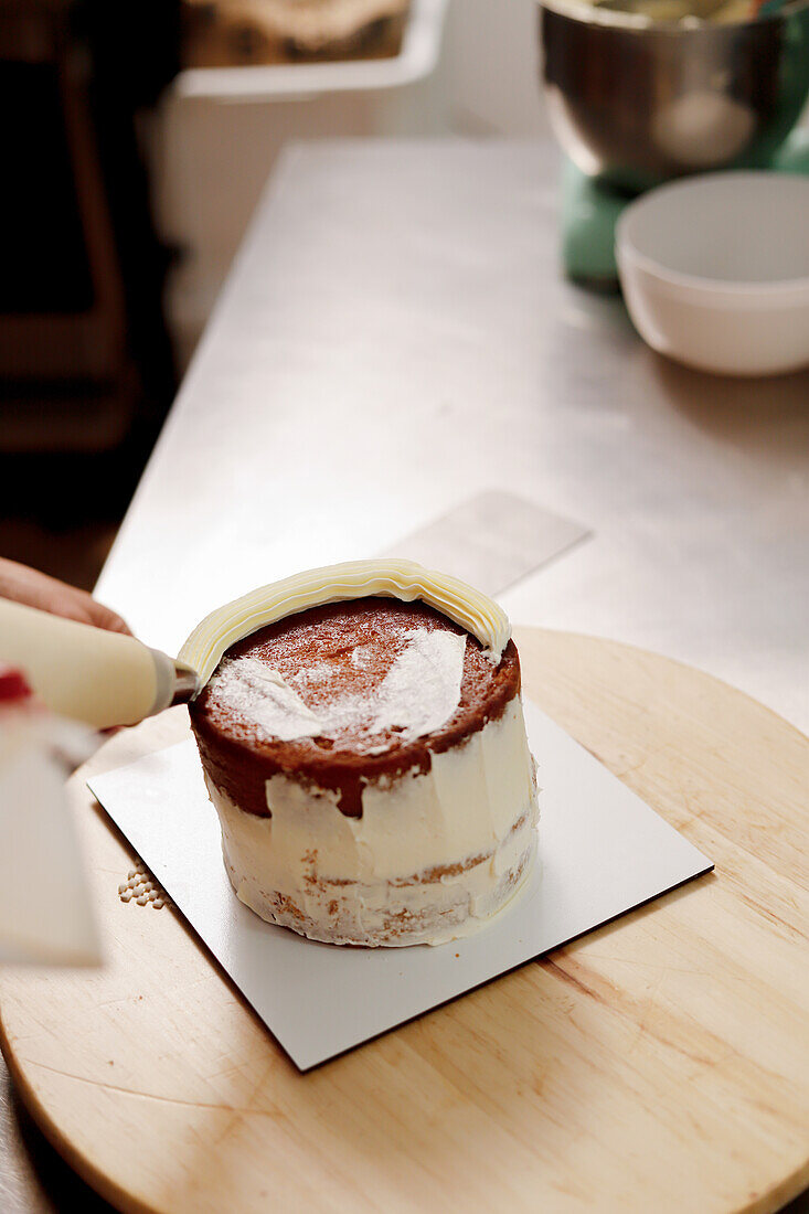 A pastry chef decorates a freshly baked cake with white icing sugar using a piping bag on a kitchen countertop, reflecting the art of cake making