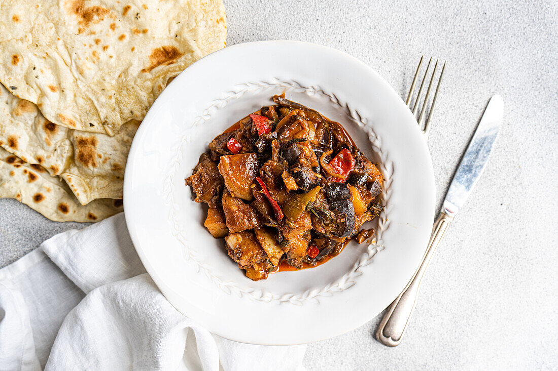 Ein schmackhafter Gemüseeintopf mit Auberginen, Tomaten, Kartoffeln, Paprika, Zwiebeln, Knoblauch und Basilikum, serviert mit hausgemachtem Fladenbrot auf einem weißen Teller