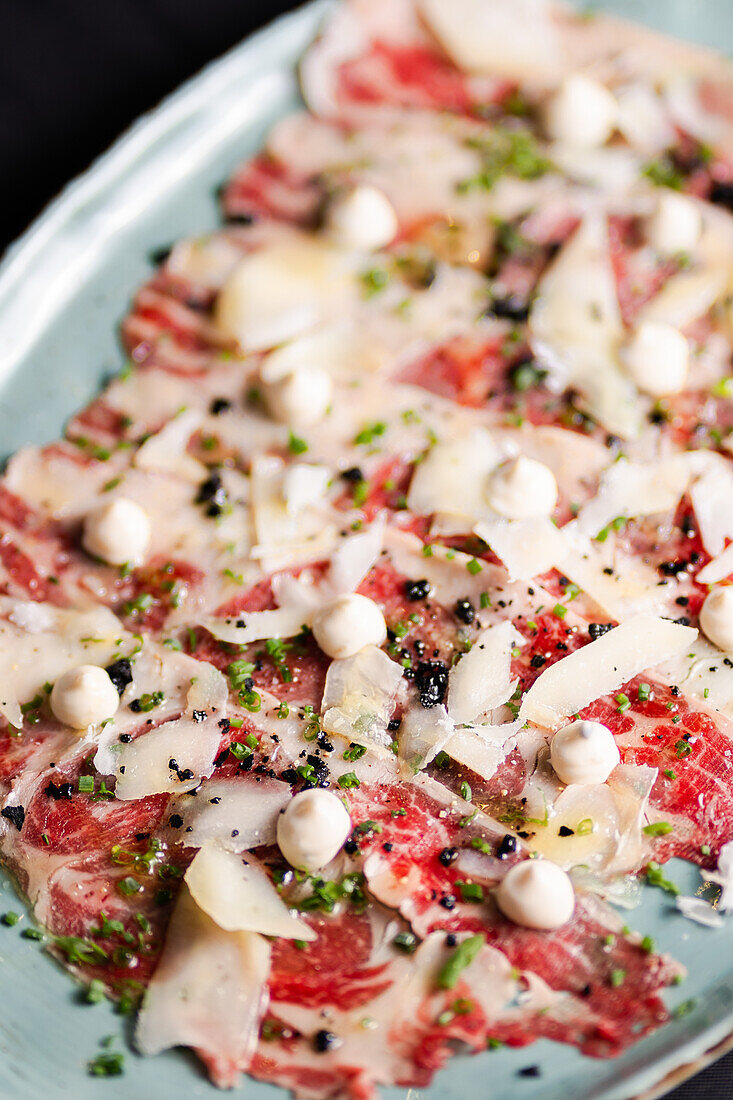 A deliciously prepared beef carpaccio garnished with shaved parmesan, fresh herbs, and seasoning, presented artistically on a plate.