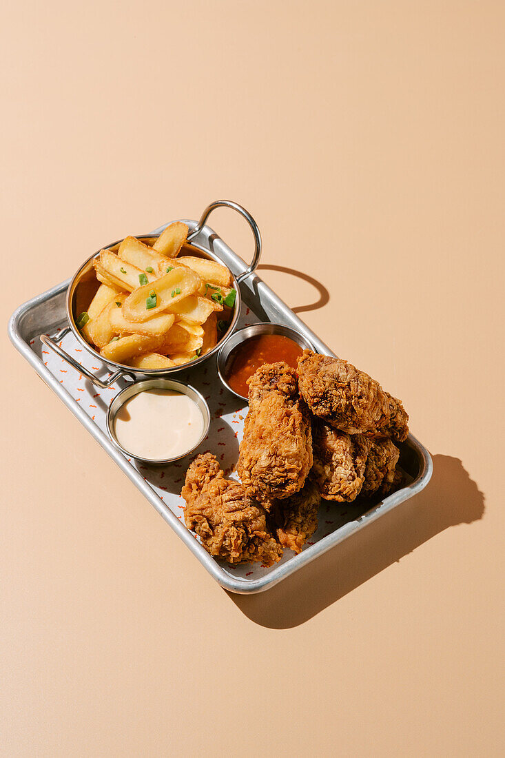 Ein Tablett mit goldbraun gebratenen Hähnchenflügeln und gewürzten Pommes frites, komplett mit cremigen und würzigen Dip-Saucen, präsentiert auf einem beigen Hintergrund.