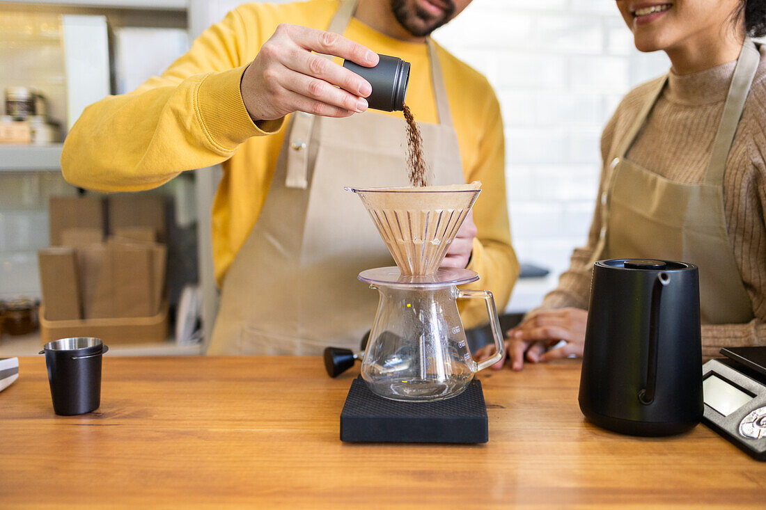 A barista in a yellow sweater pours ground coffee into a pour-over maker while colleague watches with interest, in a cozy cafe setting.