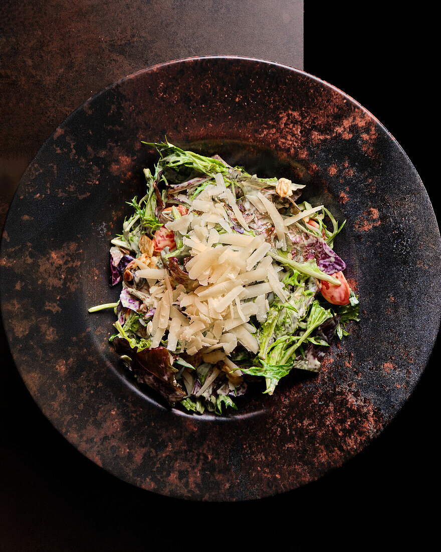 A delicious gourmet salad topped with shaved cheese, served on a textured, rustic-style plate against a dark background, perfect for a healthy meal