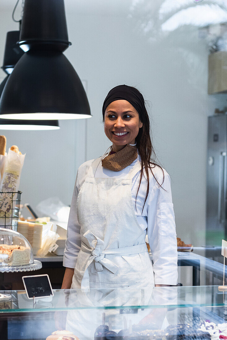 Content female baker in uniform standing behind showcase in bakehouse and looking away