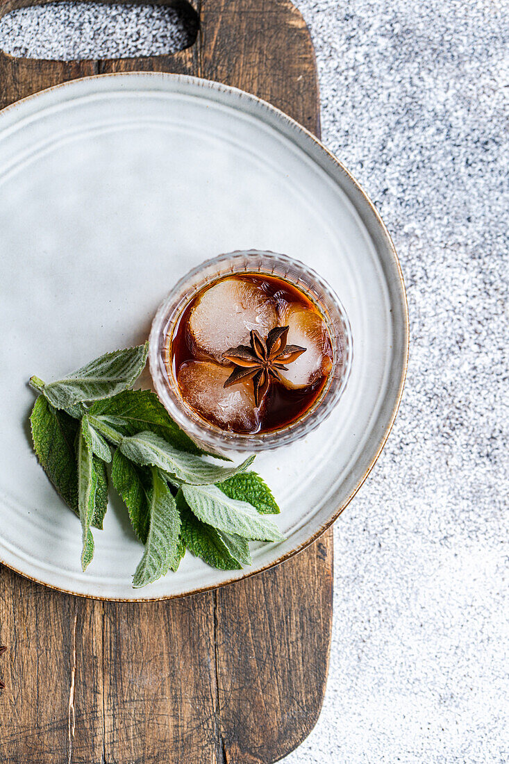 Draufsicht auf ein Glas Kirschcocktail mit Eiswürfeln und einem Zweig Minze auf einem rustikalen Servierbrett mit weißem Teller.