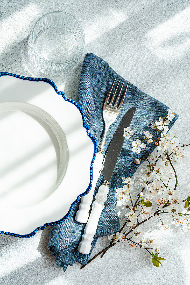 An overhead view of a stylish table setting that includes a white plate with blue rim, silver cutlery, a glass, a denim napkin, and delicate cherry blossoms