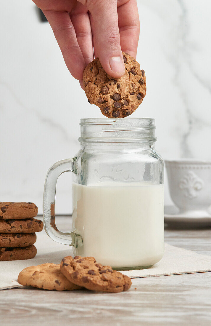 Eine Hand, die mitten in der Bewegung aufgenommen wird, als sie sich darauf vorbereitet, einen Schokoladenkeks in ein Glas mit frischer Milch zu tunken, mit weiteren Keksen und einem weißen Hintergrund