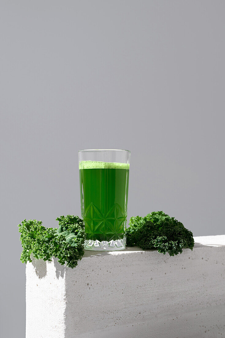 A vivid green kale juice in a transparent glass, accompanied by fresh kale leaves on a minimalist white surface, showcasing a healthy beverage option