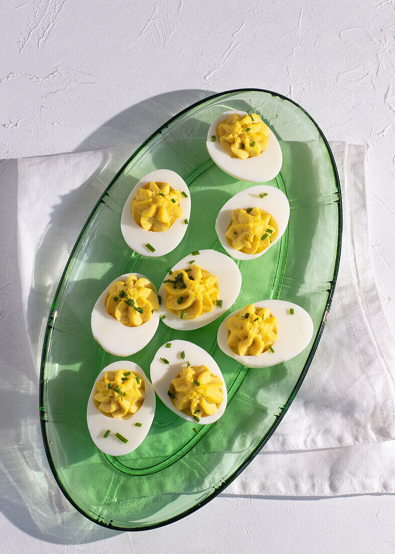 Top view of six deviled eggs neatly arranged on a green glass platter, highlighted by natural sunlight and shadows on a textured white background. Each egg is garnished with finely chopped chives.