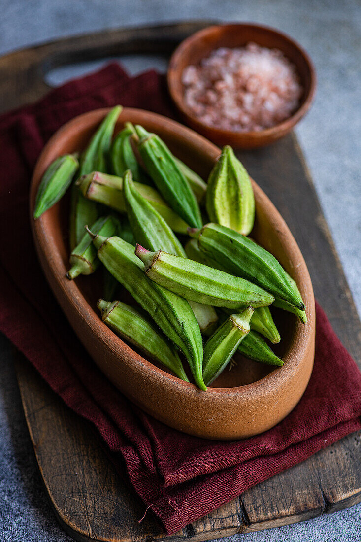 Fresh, uncooked okra bamia pods in a traditional wooden bowl, paired with a clay saucer of pink Himalayan salt, on a rustic kitchen setup