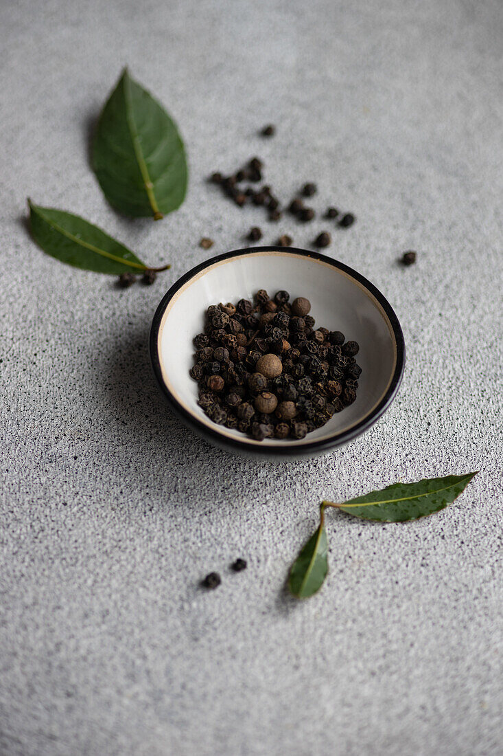 A bowl of aromatic black peppercorns accompanied by fresh bay leaves on a textured gray background.