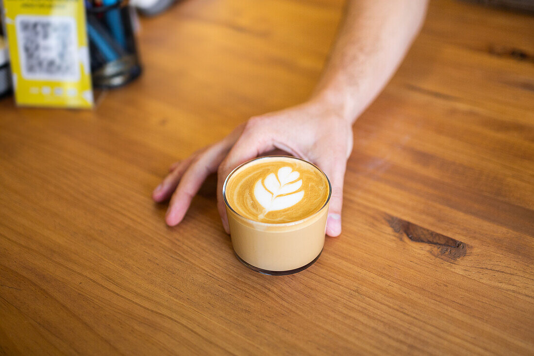 A person's hand gently grips a cup of latte with a foam art design on a wooden surface, portraying a relaxed coffee moment.