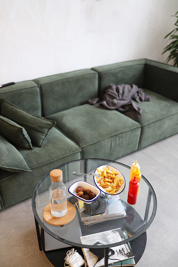 From above cozy home interior featuring a green sofa with a relaxed setup, including french fries and meatballs on a glass table. The serene ambiance is complemented by modern decor, a plant, and a throw blanket.