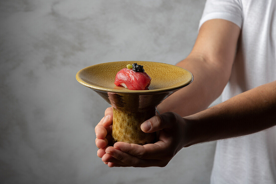 A close-up image of a male chef presenting a singular piece of sushi, topped with caviar, served in a textured ceramic dish against a soft grey background