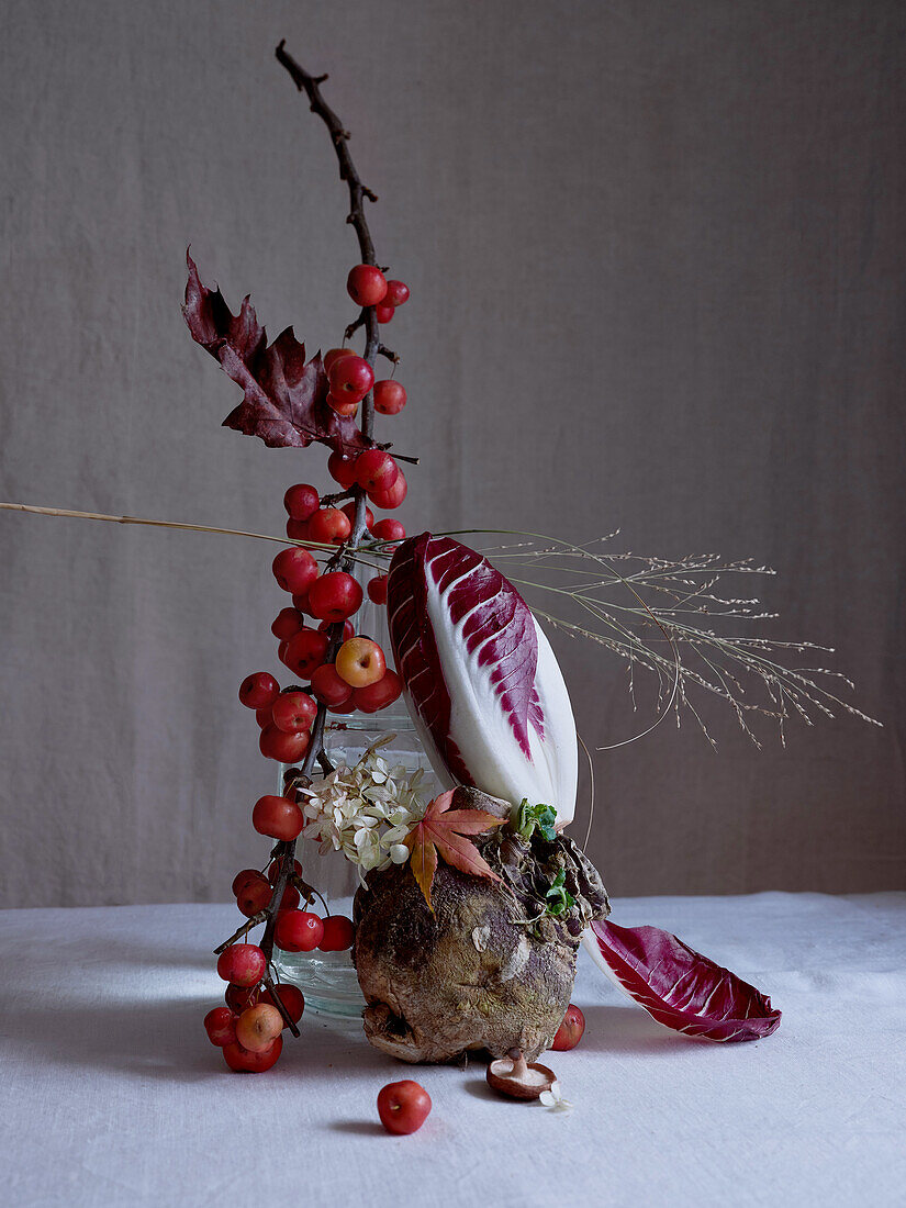 An artful arrangement featuring tiny red crab apples on branches, supported by a swede, mushrooms, radicchio leaves, and delicate white flowers, set against a soft gray backdrop.