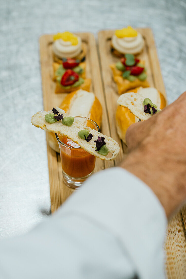 In a professional kitchen, anonymous chef deftly garnishes small glasses of soup accompanied by expertly assembled hors d'oeuvres on a wooden board