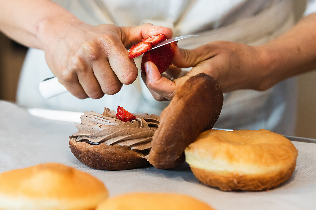 Crop faceless cook cutting fresh strawberries for decoration of sponge cake with whipped cream filling in bakehouse