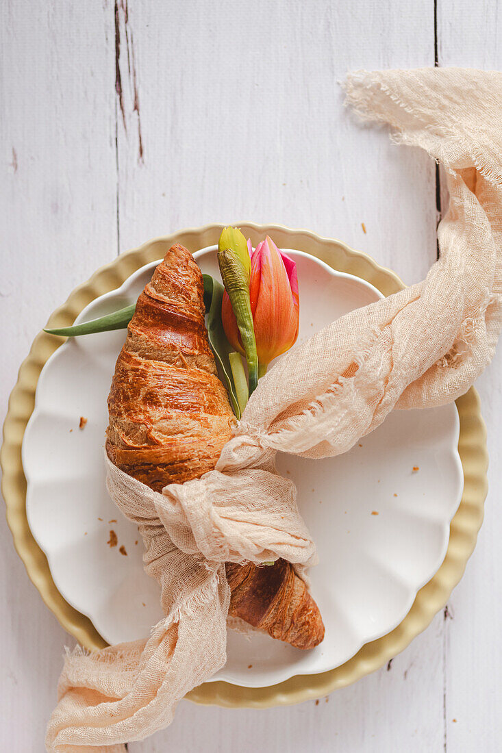 Ein mit einem rustikalen Tuch umwickeltes Oster-Croissant, verziert mit einer leuchtenden Tulpe, präsentiert auf einem dekorativen Teller, der die Essenz des Frühlings beschwört