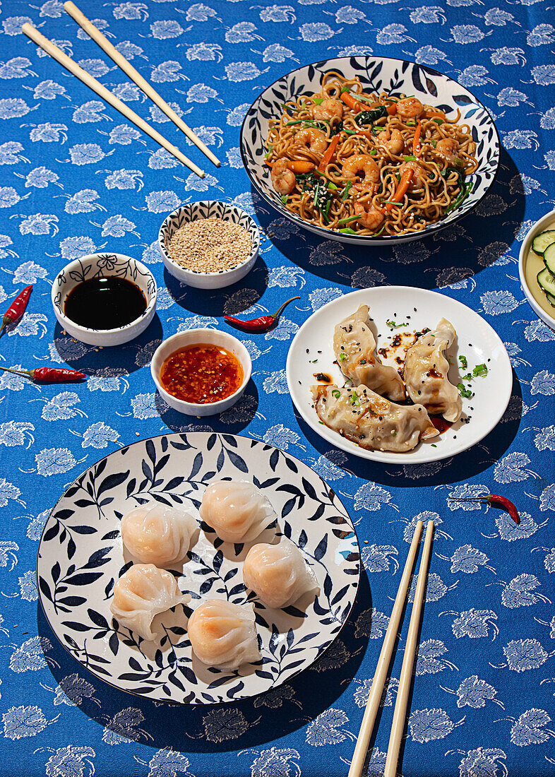 From above, a vibrant display of various traditional Chinese dishes served on a blue patterned tablecloth. Includes dim sum, noodles with shrimp, and accompanying sauces