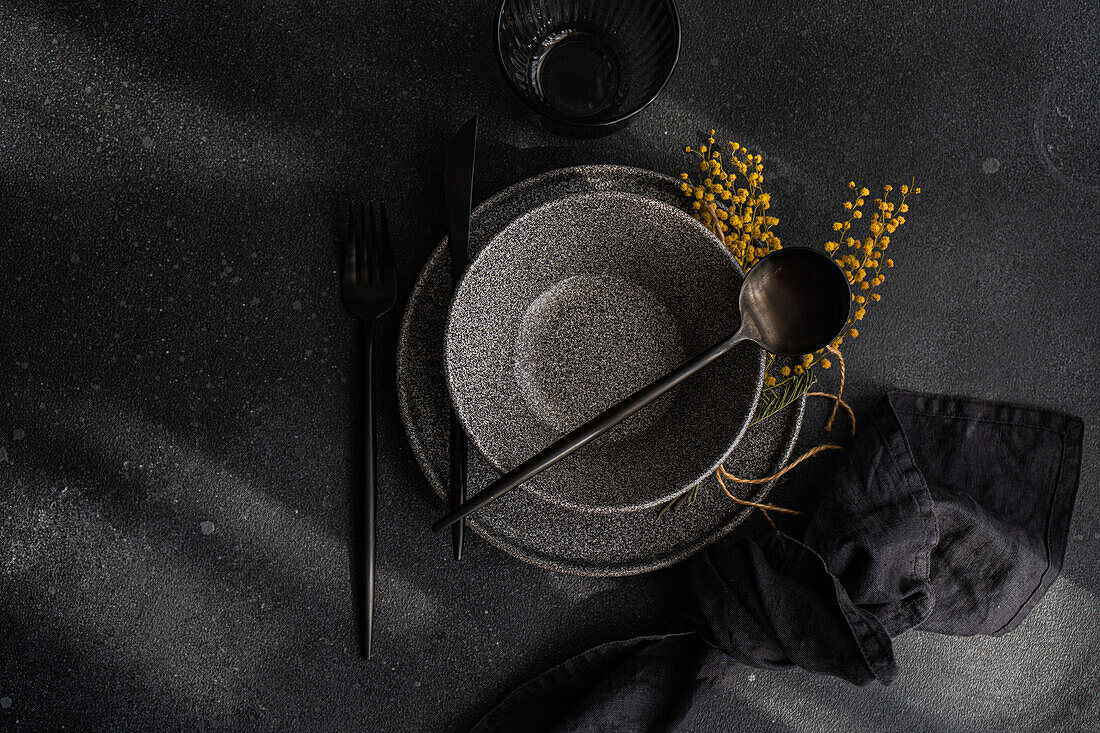 Top view of a sophisticated black table setting featuring dark plates, utensils, and a yellow mimosa garnish.