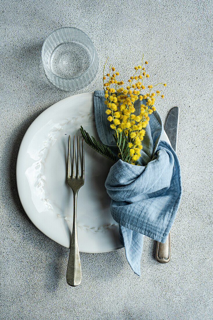 Top view of fresh outdoor table setting featuring a white embossed plate, silver cutlery, a blue linen napkin, and a sprig of bright yellow mimosa flowers