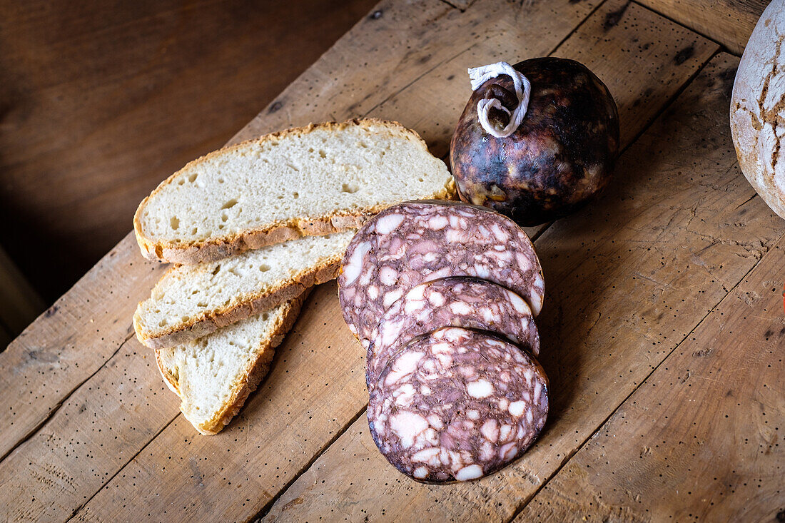 Aufgeschnittene Wurstwaren, möglicherweise Salami, und Scheiben rustikalen Brotes, die auf einem Holzbrett präsentiert werden und eine traditionelle und appetitanregende Anordnung darstellen.