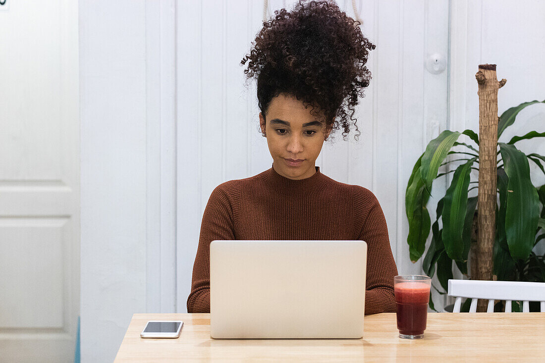 Konzentrierte afroamerikanische Freiberuflerin, die mit einem erfrischenden Smoothie am Tisch sitzt und auf ihrem Netbook surft, während sie in einem Café an einem Projekt arbeitet