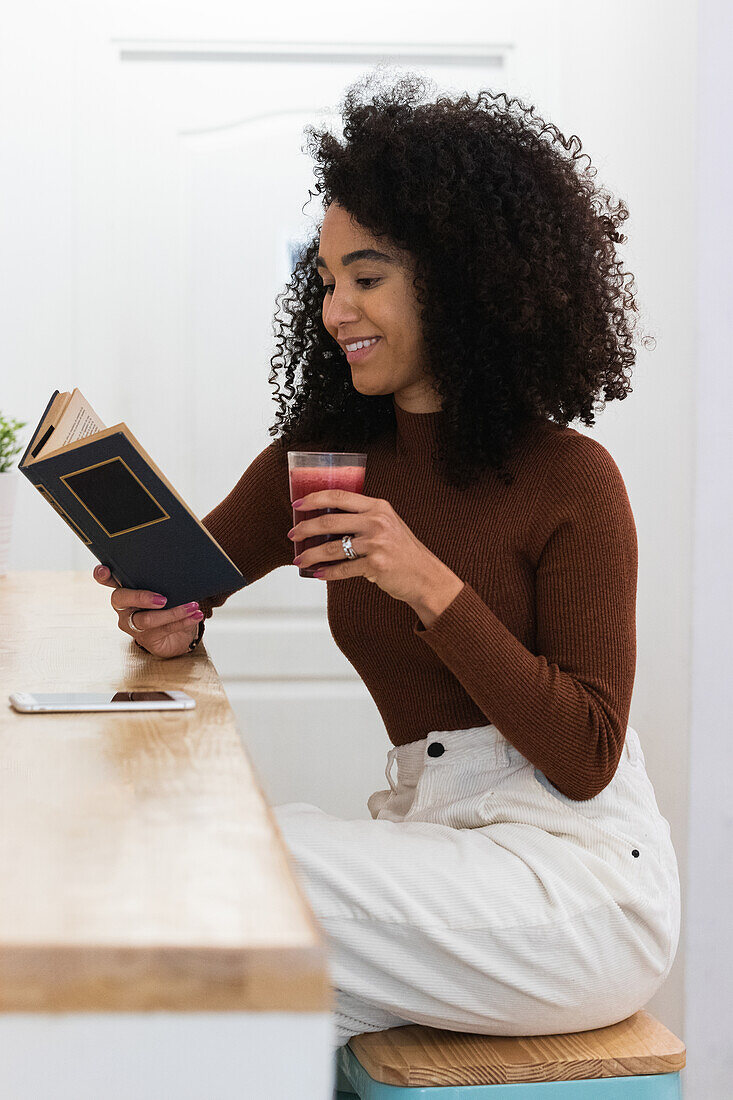 Seitenansicht einer lächelnden schwarzen Frau, die am Tresen einer Bar sitzt, ein Buch liest und einen erfrischenden Cocktail trinkt