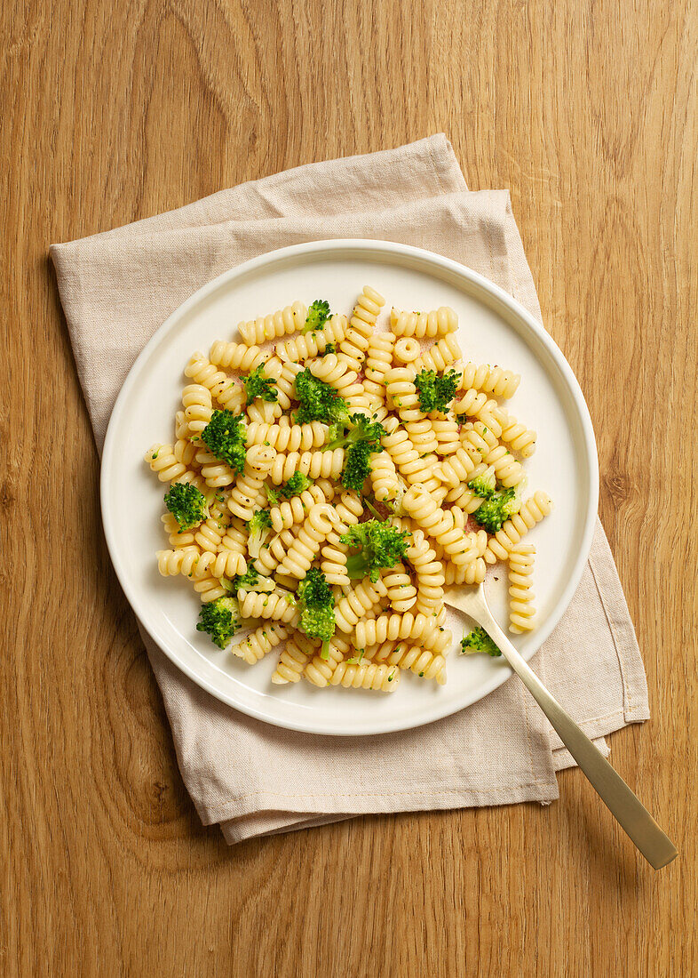 Draufsicht auf einen Teller mit gekochten Fusilli-Nudeln und frischen Brokkoliröschen, serviert auf einem Holztisch mit einer Leinenserviette