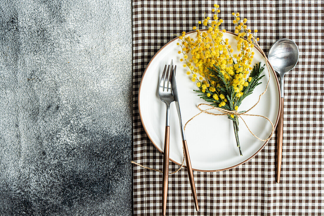 From above of white plate with a border detail, elegantly set with copper-toned cutlery and adorned with a cheerful mimosa flower arrangement, atop a checkered napkin