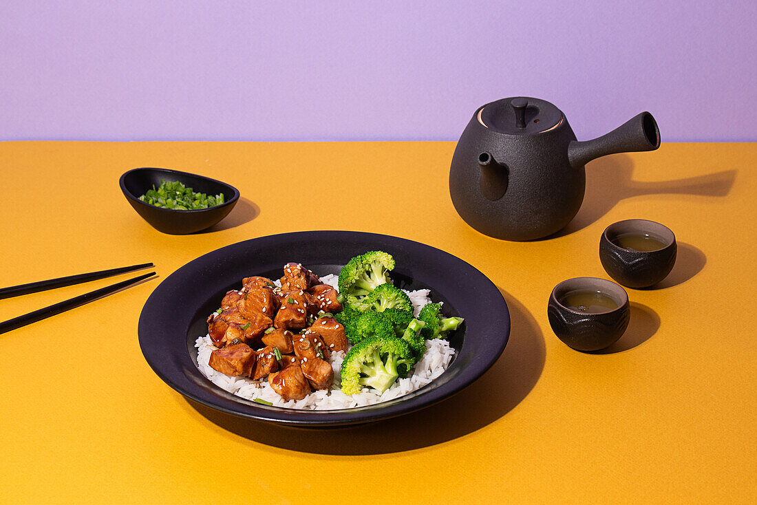 Artfully plated teriyaki chicken on a black plate, served with broccoli and rice, accompanied by a traditional tea set on a vibrant yellow and purple background.