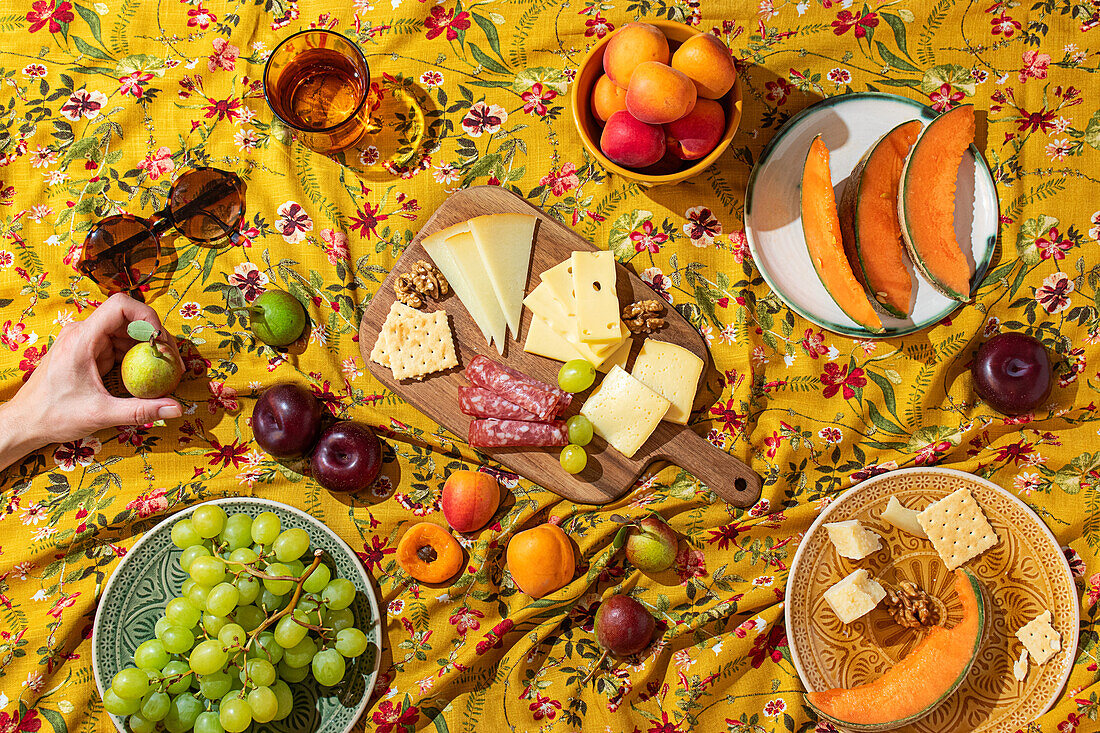 Aerial view of an anonymous hand in a colourful summer picnic setting, with a wooden cheese board laden with various cheeses, crackers and salami, surrounded by fresh fruit such as grapes, apples and melons on a flowery yellow blanket