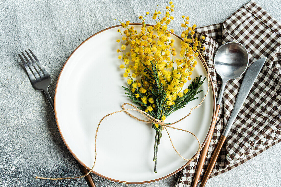 Draufsicht auf einen rustikal-schick gedeckten Tisch mit einem zarten, mit Schnur gebundenen Mimosenblumenstrauß, ergänzt durch elegantes Besteck und eine karierte Serviette