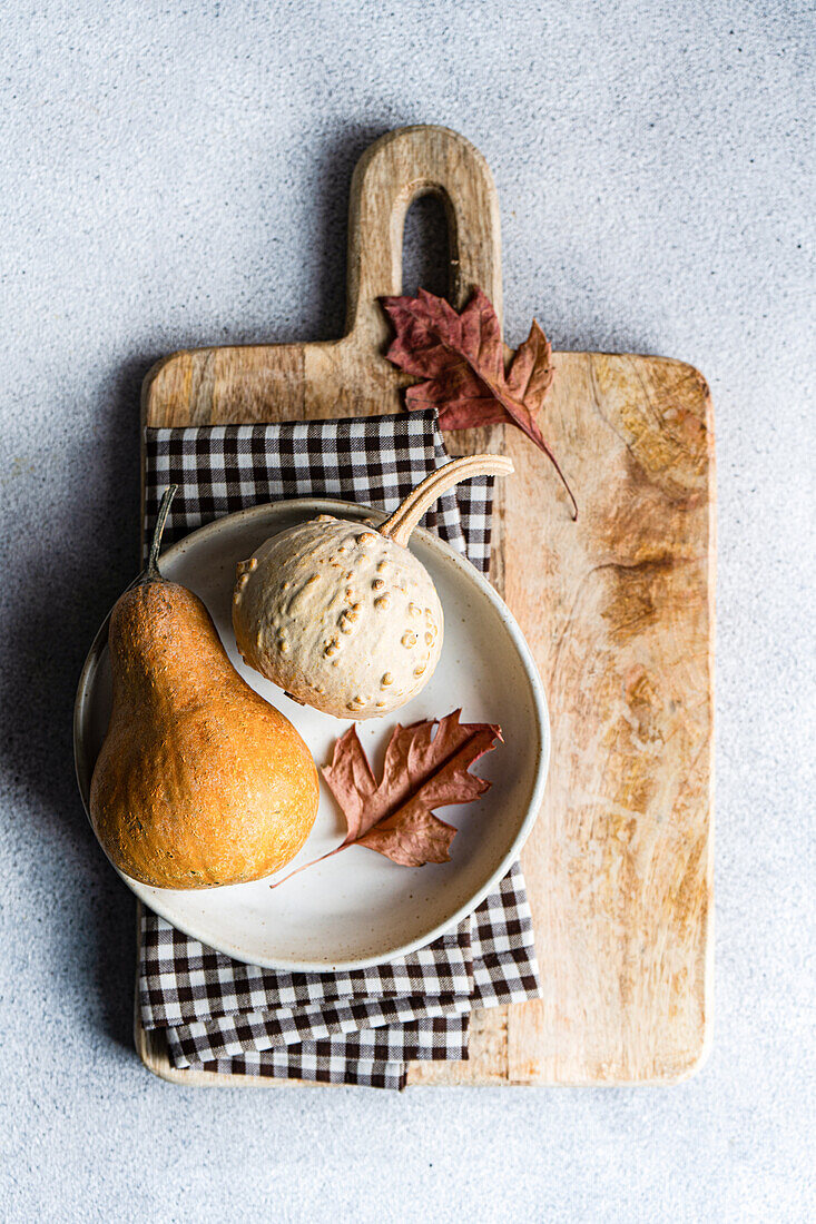 A rustic flat lay featuring a pumpkin and a gourd on a ceramic plate, placed atop a wooden cutting board with a checkered napkin and autumn leaves