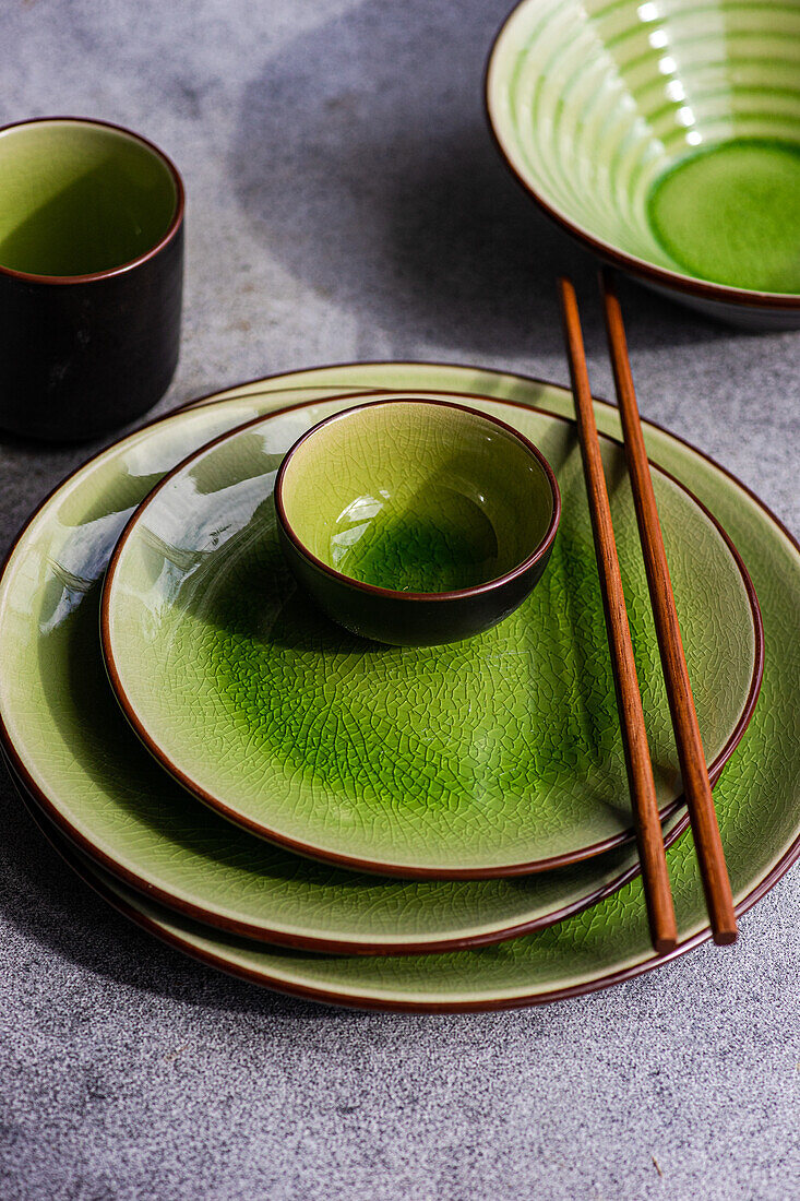 From above view of a stylish table setting featuring bright green ceramic plates and bowls, complemented by wooden chopsticks, all arranged on a textured gray surface.