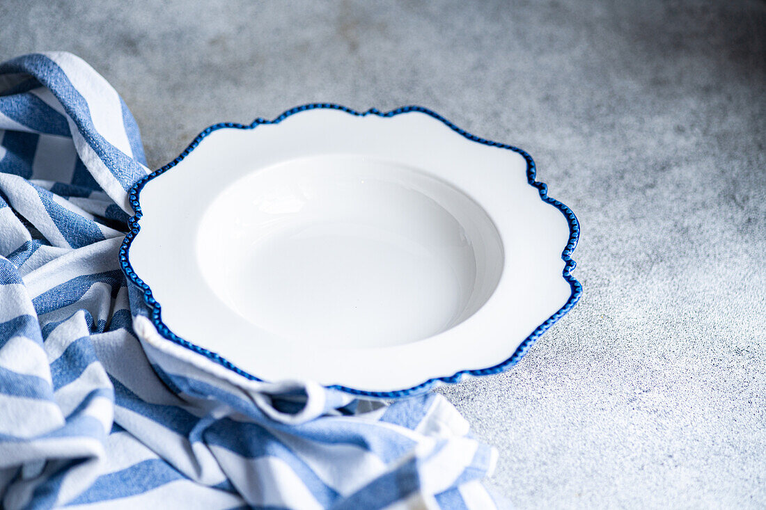 A pristine white plate with a scalloped blue edge rests on a striped cloth, presented on a textured grey surface