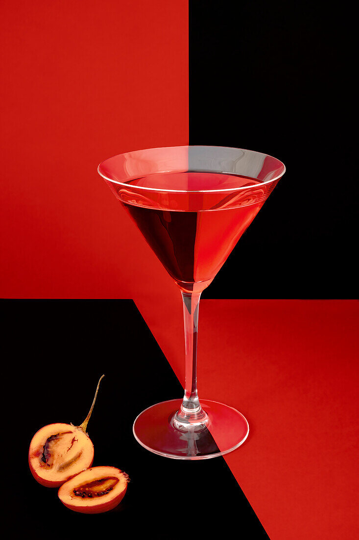 Ein leuchtend roter Cocktail in einem Martiniglas, kontrastiert mit einem gespaltenen Pfirsich auf einem zweifarbigen roten und schwarzen Hintergrund