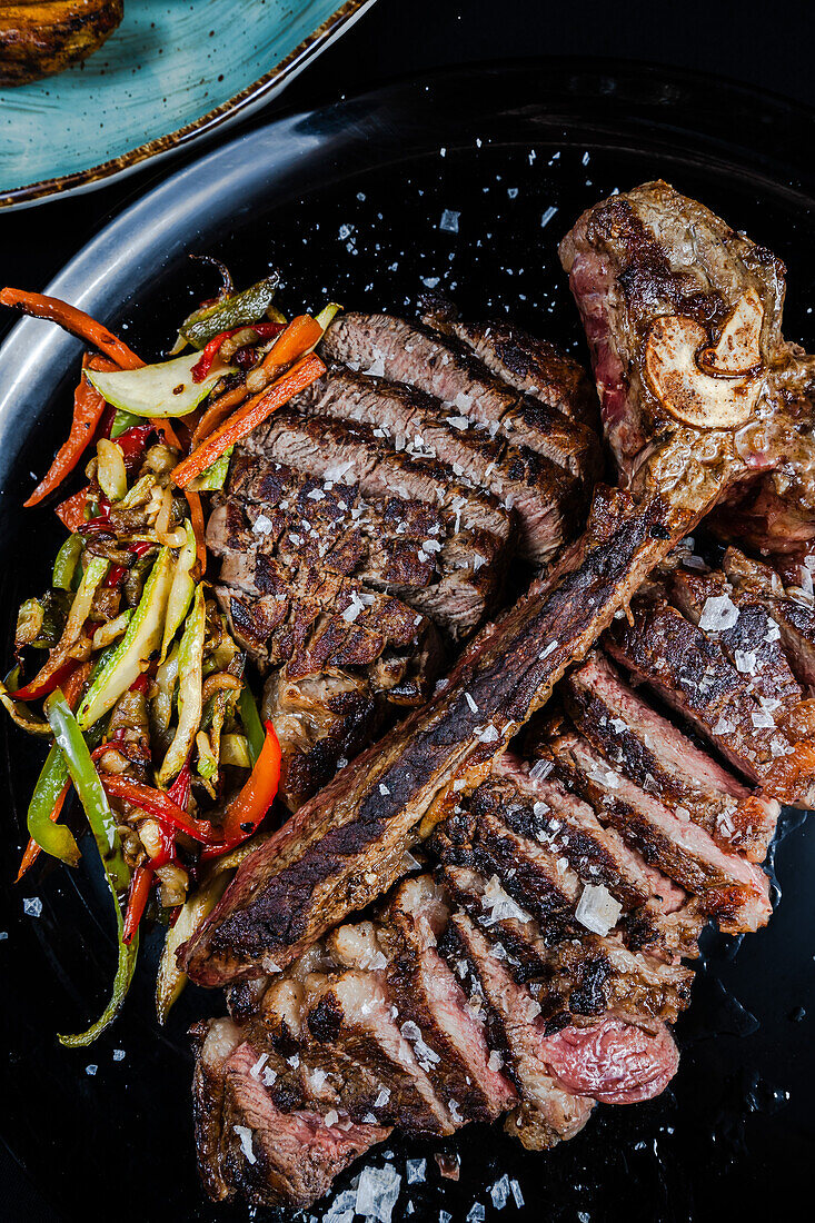 A mouth-watering 21-day aged beef T-bone steak presented with a side of colorful sauteed vegetables, garnished with flakes of sea salt.